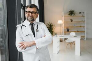 feliz confiante barbudo velho profissional médico de pé. sorridente Senior adulto médico, confiável bem sucedido terapeuta vestindo branco laboratório casaco e estetoscópio, retrato foto