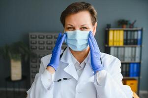 Mais velho fêmea médico vestindo face mascarar e branco médico casaco em pé dentro hospital. retrato foto