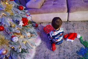 uma alegre pequeno Garoto de a Natal árvore. uma pequeno Garoto sentado em a chão Próximo para uma Natal árvore foto