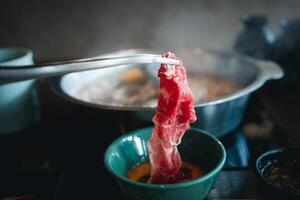 mão usar pinças com cru carne mergulho dentro cru ovo, japonês quente Panela estilo. foto