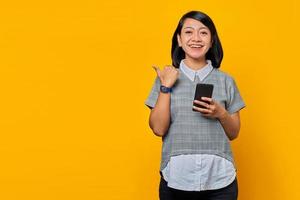 mulher asiática jovem e alegre segurando o telefone celular e apontando o dedo de lado para o espaço da cópia isolado sobre fundo amarelo