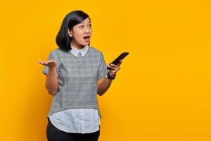 retrato de uma mulher asiática surpresa segurando um telefone celular com uma expressão confusa e infeliz em fundo amarelo foto