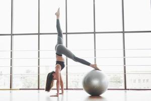 esportes é uma bela mulher que está fazendo aulas de ioga, esticando as pernas sobre a bola perto da grande janela foto