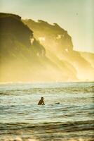 silhueta do uma surfista esperando para uma grande onda sentado em a borda às alvorecer foto