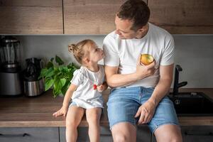 fofa pequeno menina e dela bonito Papai estão comendo fruta dentro moderno cozinha. saudável comendo. foto