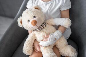 pequeno menina com quebrado dedo detém Urso de pelúcia Urso com uma enfaixado pata às a médico compromisso dentro a hospital foto