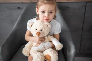 pequeno menina com quebrado dedo detém Urso de pelúcia Urso com uma enfaixado pata às a médico compromisso dentro a hospital foto