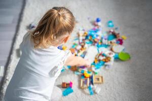 pequeno menina jogar com construtor brinquedo em chão dentro casa vivo sala, educacional jogo, gastos lazer Atividades Tempo conceito. foco em criança foto