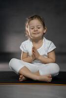 três anos velho pequeno menina meditando dentro uma lótus pose em uma cinzento fundo dentro Sombrio quarto foto