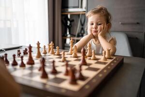 pequeno menina jogando xadrez com dela pai às a mesa dentro casa cozinha. a conceito cedo infância desenvolvimento e Educação. família lazer, comunicação e lazer. foto