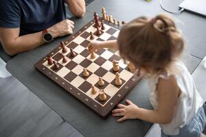 pequeno menina jogando xadrez com dela pai às a mesa dentro casa cozinha. a conceito cedo infância desenvolvimento e Educação. família lazer, comunicação e lazer. foto