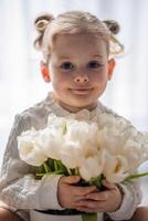 pequeno menina sentado de janela com tulipa flores ramalhete. feliz criança, dentro de casa. mãe dia, dia dos namorados dia ou aniversário conceito. foto