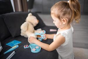 pequeno menina jogando médico com brinquedos e Urso de pelúcia Urso em a sofá dentro vivo quarto às casa foto
