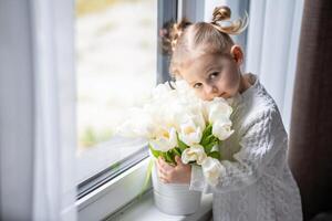 pequeno menina sentado de janela com tulipa flores ramalhete. feliz criança, dentro de casa. mãe dia, dia dos namorados dia ou aniversário conceito. foto