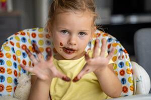 pequeno menina com loiro cabelo comendo caseiro chocolate e mostrando boca e sujo mãos com manchas do chocolate dentro casa cozinha foto