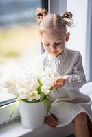 pequeno menina sentado de janela com tulipa flores ramalhete. feliz criança, dentro de casa. mãe dia, dia dos namorados dia ou aniversário conceito. foto