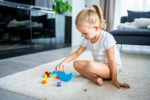 pequeno menina jogando com de madeira balanceamento brinquedo em a chão dentro casa vivo sala. foto