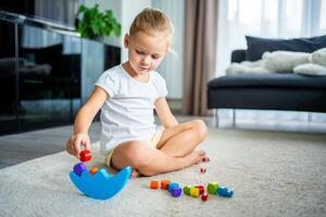 pequeno menina jogando com de madeira balanceamento brinquedo em a chão dentro casa vivo sala. foto