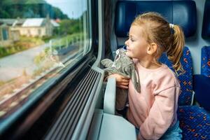 sorridente pequeno menina com brinquedo olhando Fora trem janela fora, enquanto isto em movimento. viajando de estrada de ferro, Europa foto