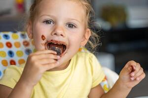 pequeno menina com loiro cabelo comendo caseiro chocolate com sujo boca e mãos dentro casa cozinha foto