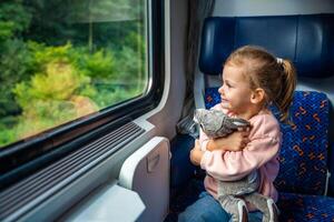 lindo pequeno menina com brinquedo olhando Fora trem janela fora, enquanto isto em movimento. viajando de estrada de ferro, Europa foto