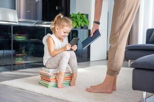 pequeno menina senta em uma pilha do crianças livros e tocam com dela Smartphone enquanto mãe dá dela uma livro foto