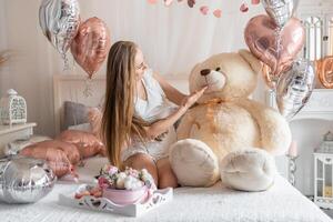 lindo jovem menina às casa em a cama dentro a manhã apreciar dia dos namorados dia celebração com coração em forma balões. Alto qualidade foto