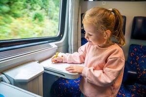 lindo pequeno menina desenha com lápis em branco papel enquanto viajando de estrada de ferro, Europa foto