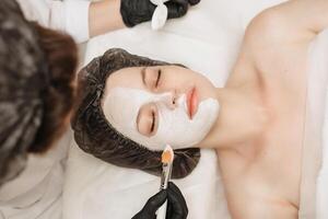 Visão a partir de acima do fêmea profissional cosmetologistas mãos aplicando mascarar para pacientes face para facial pele Cuidado. foto
