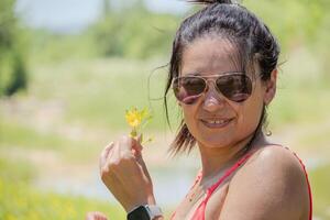 argentina mulher posando com uma flor dentro dela mão, ela desgasta óculos em uma lindo verde Prado. foto