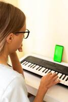 jovem mulher sentado às musical teclado usando Smartphone com verde tela para estudando jogando piano foto