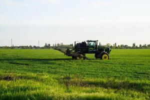 trator com a Socorro do uma pulverizador sprays líquido fertilizantes em jovem trigo dentro a campo. foto