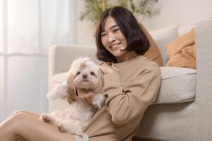 feliz jovem ásia mulher abraço e gastos Tempo com fofa cachorro dentro vivo sala. foto