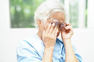 ásia Senior mulher vestindo Óculos reduzir dor de cabeça e fadiga ocular Sofra às casa Cuidado serviço. foto