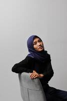 sério ásia hijab mulher dentro casual camisa sentado em cadeira, posando com vários mão gesto. isolado estúdio retrato em cinzento fundo. foto