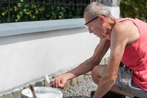 uma grisalho meia idade homem picaretas acima branco pintura a partir de uma balde com uma escova para pintura a cerca foto