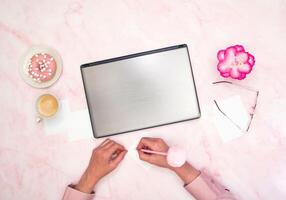 escritório trabalhador dentro uma Rosa camisa senta às uma computador portátil, topo ver, próximo para uma copo do café foto