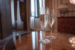 elegante champanhe flautas em Castanho mesa dentro luxo hotel quarto com lustre fundo foto
