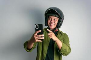adulto ásia homem sorridente quando olhando para dele handphone enquanto vestindo motocicleta capacete foto