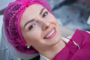 retrato do jovem sorridente mulher sentado dentro estomatologia clínica cadeira e médico mãos com dental boca espelho e dental explorador Ferramentas preparando para examinar dentes doença. foto