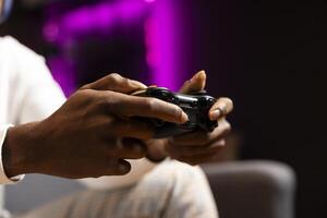 fechar acima tiro do controlador aguarde de jogador às casa jogos e desfrutando lazer tempo. africano americano homem usando controle dentro Rosa néon aceso vivo sala, jogando videogame em console, relaxante foto