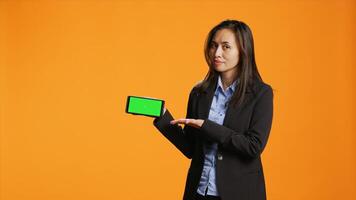 empresária segurando telefone com tela verde modelo, posando contra laranja fundo e mostrando em branco copyspace dentro estúdio. ásia adulto com chromakey tela em Smartphone aplicativo. foto