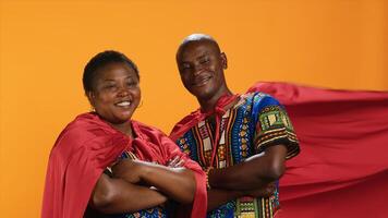 africano americano casal posando com Super heroi vermelho capa, agindo Como poderoso sério Heróis com quadrinho livro mantos. étnico pessoas em pé dentro estúdio com braços cruzado, orgulhoso Forte modelos. foto