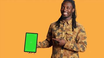africano americano homem apresenta tela verde em tábua, mostrando isolado copyspace tela e sorridente dentro frente do Câmera. jovem pessoa segurando Móvel gadget com em branco brincar exibição layout. foto
