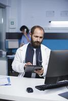 usando uma tábua, masculino médico lê paciente registros. caucasiano homem dentro uma laboratório casaco é revendo médico dados em uma Área de Trabalho computador e digital dispositivo dentro a clínica escritório. foto