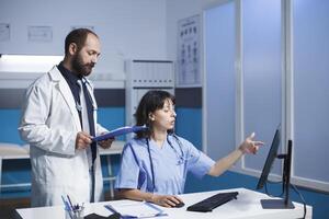 cuidados de saúde trabalhadores discutir médico resultados e tratamento opções usando tecnologia dentro clínica sala. imagem mostra enfermeira dentro azul esfrega indicando paciente em formação em pc monitor para médico com tábua. foto