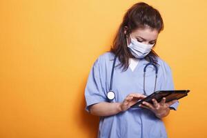 fêmea médico segurando digital dispositivo para digitando paciente dados enquanto vestido dentro azul esfrega e cobiçado proteção mascarar dentro estúdio. médico com estetoscópio usando tábua contra isolado fundo. foto