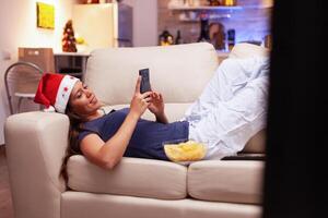 mulher deitado em sofá navegando em social meios de comunicação usando Smartphone durante época de Natal dentro natal decorado cozinha. menina desfrutando inverno feriado a comemorar Natal estação às casa foto