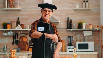 positivo chefe de cozinha gravação dentro cozinha uma Novo receita para vídeo canal. aposentado blogueiro padeiro influenciador usando Internet tecnologia comunicar, tiroteio, blogging em social meios de comunicação com digital equipamento foto