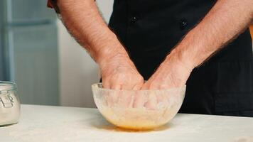 Senior chefe de cozinha aspersão farinha e amassar massa dentro vidro tigela. fechar aposentado idosos padeiro com bonete e avental, espalhando, peneiramento peneirar cru ingredientes de mão cozimento caseiro pizza, pão. foto